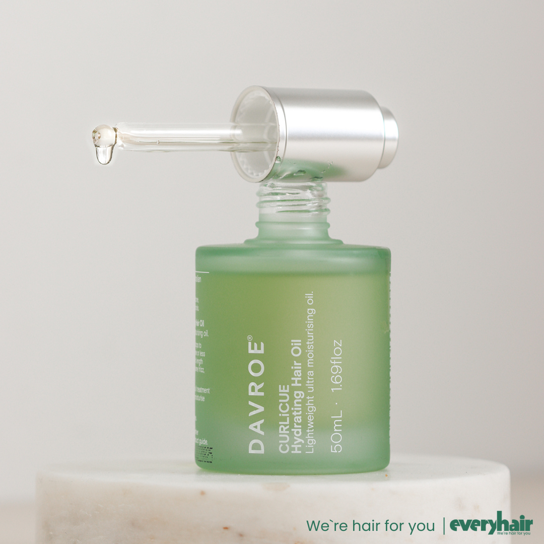 Grønn frostet DAVROE Curlicue Hydrating Hair Oil-flaske med pipette og dråpe olje, stående på lys steinpedestal mot nøytral bakgrunn.