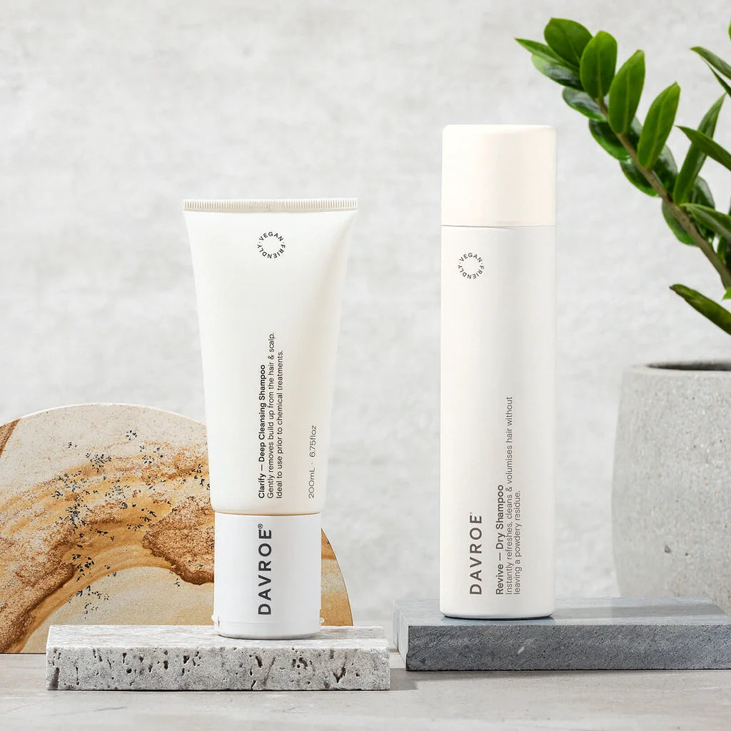 To hvite DAVROE hårpleieprodukter – en tube og en høy flaske – plassert på steinblokker foran lys bakgrunn, med grønn plante og naturstein som dekor.