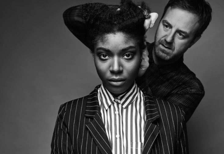 Svart-hvitt portrett av en modell med krøllete hår i stripet dressjakke, mens en frisør står bak og styler håret hennes i studio