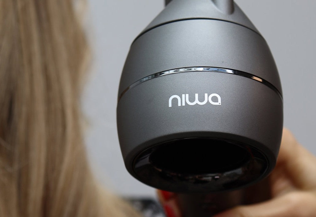 Nærbilde av grå NIWA hårføner rettet mot langt blondt hår, med tydelig niwa-logo på munnstykket.