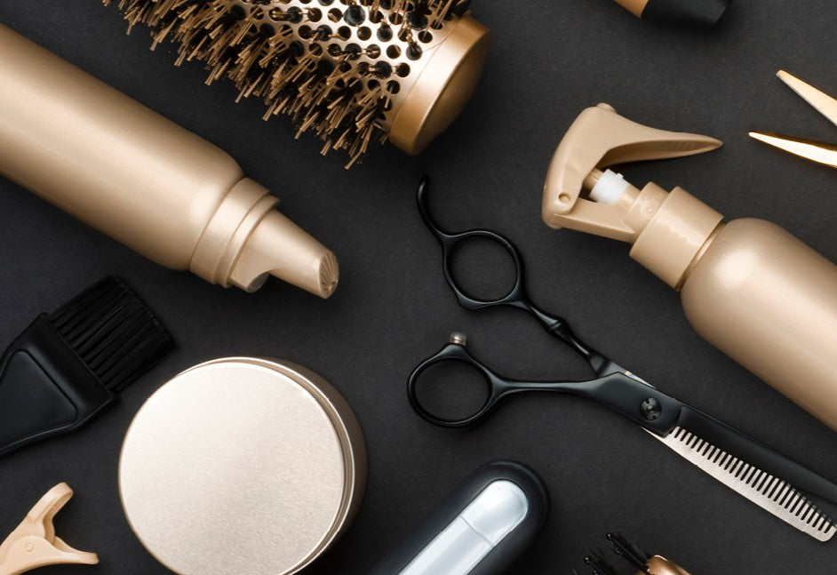Flatt lay av frisørutstyr – gullfargede flasker, rundbørste, krukker, klips og svarte sakser – arrangert på mørk bakgrunn.