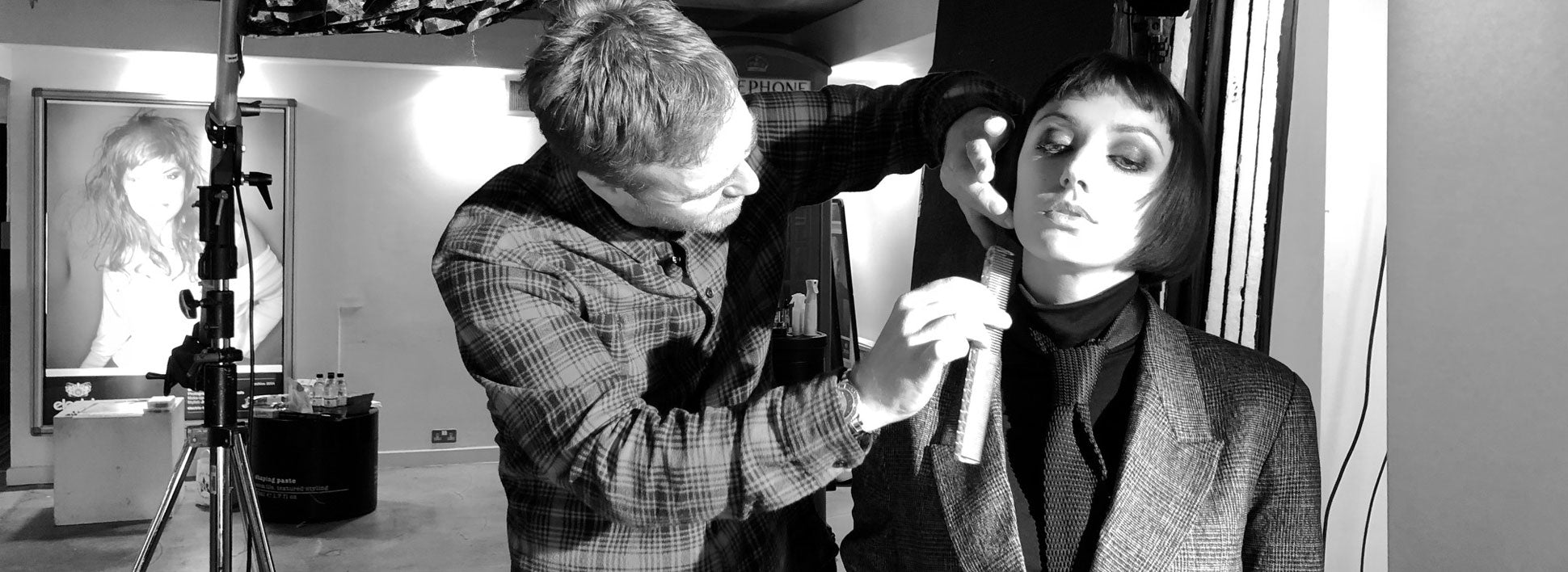 Sort-hvitt-bilde av stylist som former håret til en modell med kort bobfrisyre i fotostudio, med studioutstyr og lys i bakgrunnen.